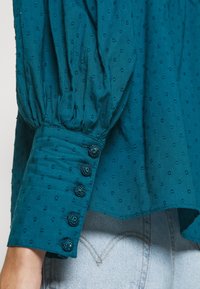 Grønblå tekstureret bluse med lange ærmer og fire stofbetrukne knapper ved manchetten, båret over lyseblå denimjeans.