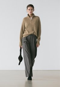 Pull en tricot beige avec un col montant et un motif torsadé, associé à un pantalon plissé gris et des chaussures noires, tenant un sac à main sombre.