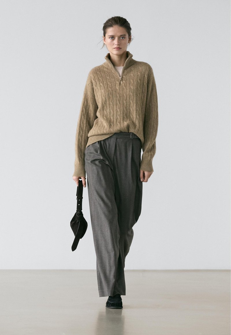 Pull en tricot beige avec un col montant et un motif torsadé, associé à un pantalon plissé gris et des chaussures noires, tenant un sac à main sombre.