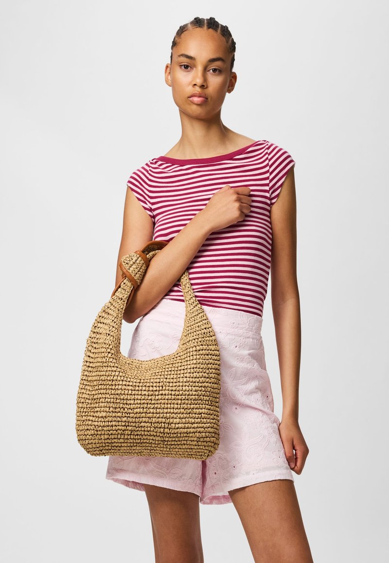 Femme aux cheveux tressés portant un haut rayé rouge et blanc et un short rose clair brodé, tenant un grand sac en paille tressée.
