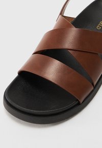 Pruun nahast sandaal ristatud rihmadega ja musta tekstuurses tallaga. Avatud tagaosa, sile viimistlus ning minimalistlik välimus.