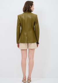 Blazer in pelle verde oliva con design aderente, cuciture delle spalle pronunciate e texture liscia, abbinato a pantaloni corti beige.