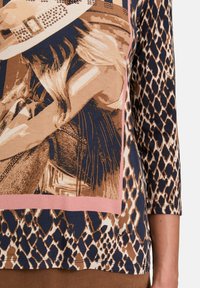 Bluzka z długim rękawem w wzór zwierzęcy, z graficznym wzorem w ziemistych kolorach, zdobieniami oraz kontrastującą różową lamówką. Miękka w dotyku.