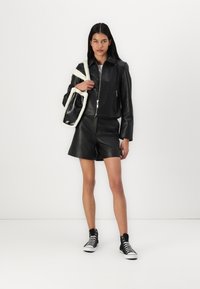 Svart lær jakke med cropped design og glidelås, kombinert med svarte lærshorts. Tilbehør inkluderer en svart og hvit pelskledd veske og sneakers.