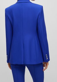 Niebieska marynarka o dopasowanym kroju, gładkim materiale i usztywnionych ramionach. Na mankietach znajdują się po cztery srebrne guziki, z tyłu marynarki jest rozcięcie.