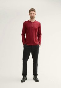 Langärmliges bordeauxrotes Henley-Hemd mit Knopfdetails, kombiniert mit schwarzen Hosen und schwarzen Schuhen. Minimalistisches Design vor hellem Hintergrund.