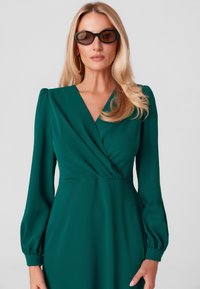 Zielona sukienka w stylu owijanym z długimi, bufiastymi rękawami i głębokim dekoltem w kształcie litery V. Gładki materiał z subtelnymi detalami drapowania. Modelka nosi czarne, owalne okulary przeciwsłoneczne.