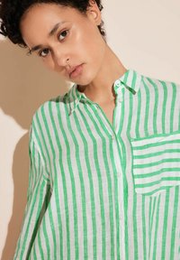 Green and white striped button-up shirt made of lightweight fabric, featuring a chest pocket and a collared neckline. Textured surface.