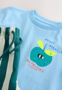Světle modrá košile s kresleným zeleným ovocem a žlutými listy, text "Fruit Market Miércoles 6:00 AM," v kombinaci se zelenými a bílými pruhovanými kalhotami.