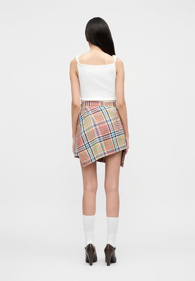 Γυναίκα που στέκεται πλάτη, φοράει λευκό αμάνικο τοπ, ασύμμετρη πολύχρωμη φούστα με καρό σχέδιο, λευκές κάλτσες και καφέ τακούνια.
