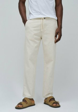 Pantalones de lino mezclado en color beige claro, con un corte relajado, cintura con cordón y bolsillos laterales, combinados con sandalias marrones con detalle de hebilla.