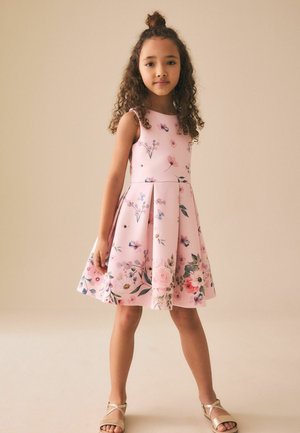 Niña joven con cabello rizado, vestida con un vestido rosa sin mangas con flores y sandalias beige, de pie contra un fondo beige liso.