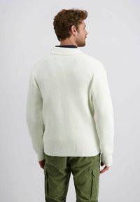 Cremfarbener Strickpullover mit strukturiertem Muster und Schalkragen, kombiniert mit grünen Cordhosen, von hinten betrachtet.