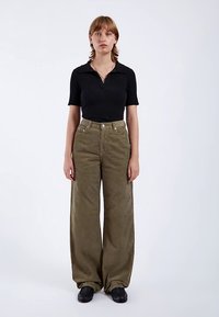 Polo de manga corta negra con costillas, pantalones de pana de pierna ancha en verde oliva, bolsillos delanteros, botones metálicos, tela texturizada y zapatos negros.