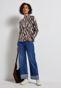 Haut à manches longues à motif zèbre en beige et marron foncé, associé à un jean en denim bleu à jambes larges, avec des poignets repliés et des baskets blanches. Sac à épaule marron.