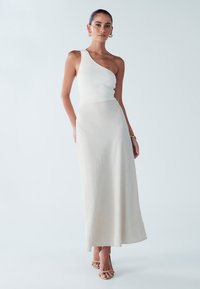 Haut asymétrique à une épaule en maille côtelée blanche, longue jupe fluide beige à texture douce. Porté avec des sandales à talons hauts couleur tabac et des boucles d'oreilles dorées.