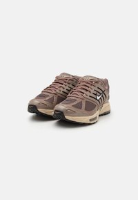 Beige Mesh-Laufschuhe mit dunklen Akzenten, strukturierten Oberflächen und einer dicken Sohle. Verfügt über ein auffälliges Logo und ein effizientes Schnürdesign.