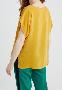 Blusa gialla a maniche corte con una texture liscia e scollo arrotondato; abbinata a pantaloni verdi con righe nere laterali.