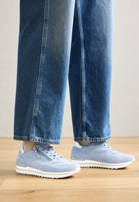 Light blue suede sneakers with white soles, featuring a zipper and lace-up design, paired with loose blue denim trousers.