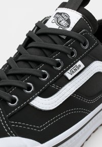 Black canvas sneaker with white accents, featuring metal eyelets, black laces, stitched details, and a Vans logo tag at the tongue.