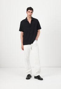 Camicia nera a maniche corte con righe verticali, chiusa con bottoni frontalmente, abbinata a pantaloni bianchi a vestibilità ampia e scarpe nere, su uno sfondo neutro.