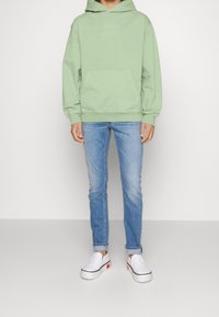 Sudadera con capucha verde menta con un bolsillo tipo canguro delantero, combinada con pantalones vaqueros azul claro y zapatos blancos sin cordones con un detalle rojo.