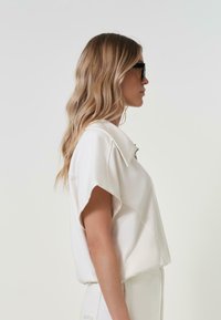 Lys beige kortermet topp med flat krage, med en avslappet passform og glatt tekstur, stylet med høyt liv og lyse bukser.