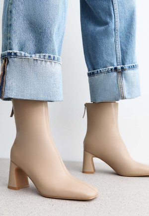 Bottines beiges en cuir lisse, avec un bout pointu et un large talon bloc, présentées aux côtés d'un jean bleu clair retroussé.