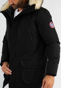 Parka noire isolée avec capuche en fourrure, poches zippées et poignets élastiques. Présente un patch logo sur la manche gauche.