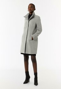 Grau wollener Mantel mit hohem Kragen, seitlichen Taschen und geradem Schnitt. Das Modell trägt ein schwarzes Kleid und Stiefeletten. Glatte Textur, maßgeschneiderter Schnitt.