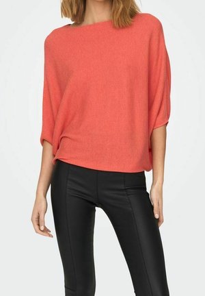 Femme portant un haut corail ample à manches courtes et un pantalon noir moulant, debout devant un fond clair uni.