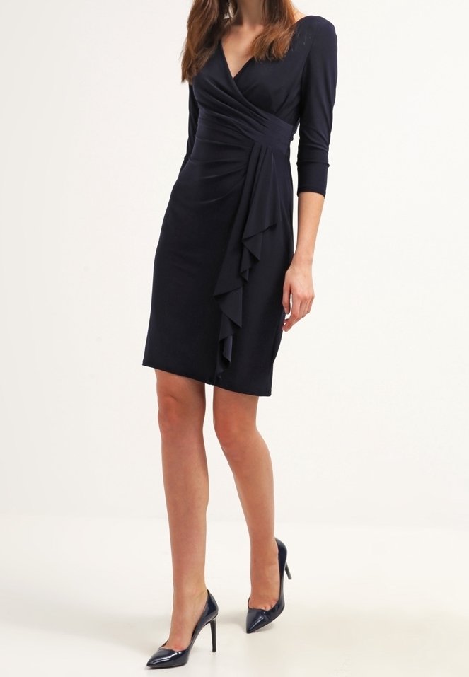 Nainen, joka on pukeutunut polvimittaisiin laivastonsinisiin kietaisumekkoihin, joissa on röyhelöyksityiskohta ja kolmanneksen pituiset hihat. Kengät ovat laivastonsinisiä, kärki korkeakorkoisia.