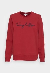 Sudadera roja con cuello redondo, que presenta un logotipo bordado en negro de "Tommy Hilfiger" en el pecho. Fabricada con una tela suave y con puños acanalados.