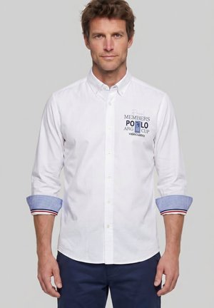 Hombre con camisa blanca de manga larga abotonada, con puños a cuadros azules y pantalones azul marino, de pie frente a un fondo gris liso.