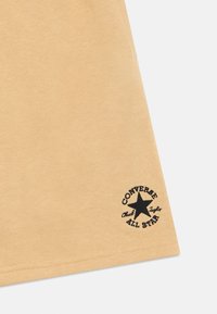 Ανοιχτό μπεζ βαμβακερό μπλουζάκι με μαύρο κυκλικό λογότυπο που περιλαμβάνει ένα αστέρι και το κείμενο "Converse Chuck Taylor All Star" στο τελείωμα.