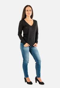 Donna in piedi con le mani nelle tasche dei jeans, indossante un top nero a maniche lunghe con scollatura a V, jeans blu e tacchi neri su uno sfondo bianco.