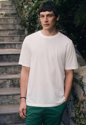 Junger Mann mit kurzen dunklen Haaren, der ein weißes T-Shirt und grüne Hose trägt und in der Nähe von Steintreppen und Grünflächen steht.