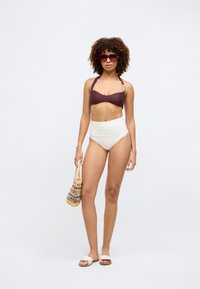 Bikini mit einem strukturierten dunkelvioletten Oberteil und einem hoch taillierten weißen Höschen. Accessoires mit einer geflochtenen Tasche und flachen Sandalen.