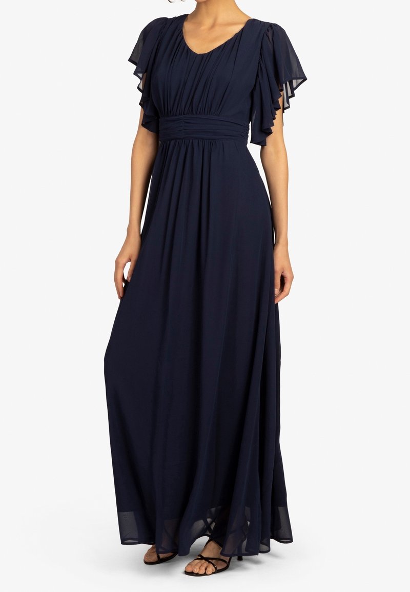 Femme portant une robe longue bleu marine avec des manches flottantes et une taille froncée, associée à des sandales noires à brides.