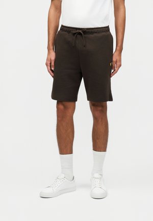 Άνδρας με σκούρα καφέ σορτς με κορδόνι, λευκές κάλτσες και λευκά sneakers, στέκεται σε ένα απλό φωτεινό φόντο.