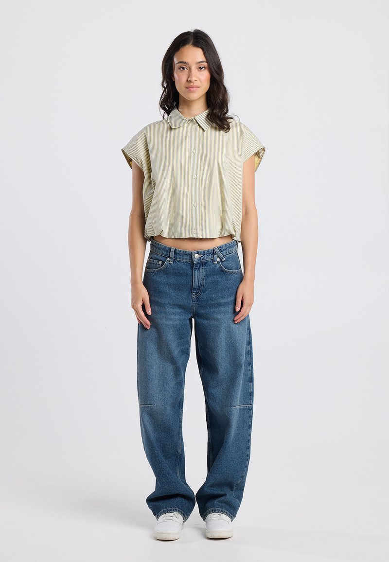Chemise courte à manches courtes de couleur jaune clair avec des rayures blanches ; associée à un jean en denim bleu foncé à coupe ample. Des sneakers blanches complètent le look.