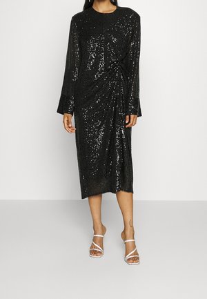 Schwarzes Midikleid mit langen Ärmeln, glänzenden Pailletten und seitlicher Raffung, getragen mit weißen, hochhackigen Riemchensandalen auf hellem Hintergrund.