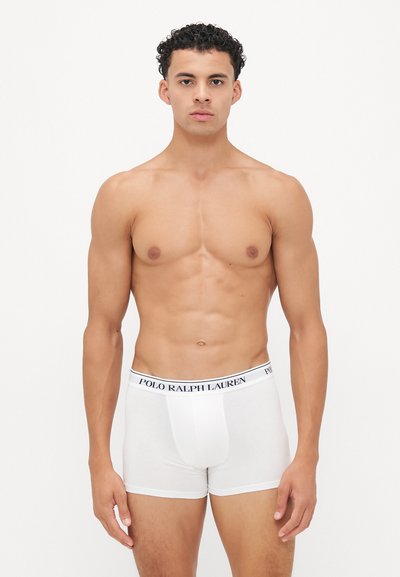 Moški model s kodrastimi lasmi stoji obračunano naprej, oblečen v bele bokserice Polo Ralph Lauren pred enobarvno belo ozadje.