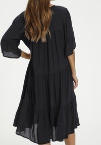 Mujer con cabello largo y ondulado, vistiendo un vestido negro suelto de volantes con mangas hasta el codo, de pie con una mano en la cadera, de espaldas.