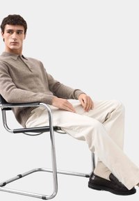 Beżowy dzianinowy sweter z kołnierzykiem, noszony na kremowych spodniach. Model siedzący na chromowanym krześle, w ciemnych butach. Proste, neutralne kolory.