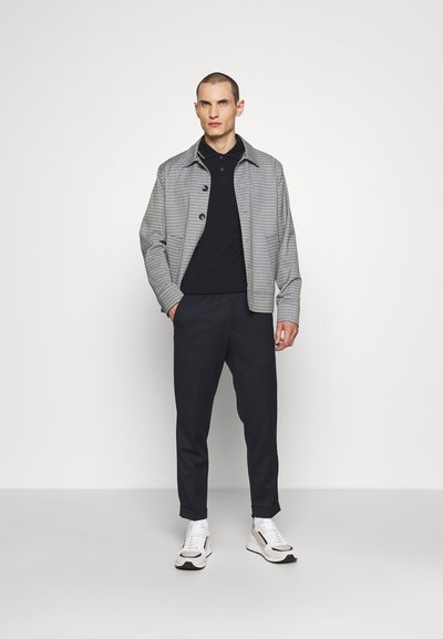 Gray checkered jacket over a black polo, paired with dark tapered pants and white sneakers. Features rounded collar and button closure.