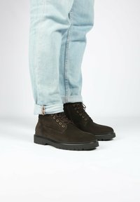 Stivali alla caviglia in suede marrone con tacco basso, punta rotonda e lacci scuri, abbinati a jeans in denim blu chiaro con risvolto. Texture liscia e suola robusta.