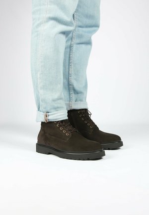 Botas de tobillo de ante marrón con un tacón bajo, punta redonda y cordones oscuros, combinadas con jeans de mezclilla de un azul claro y con dobladillo. Textura suave y suela resistente.
