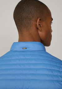 Ljusblå isolerad jacka med horisontell quiltad textur och en märkesetikett på kragen. Slätt tyg och strukturerad design.