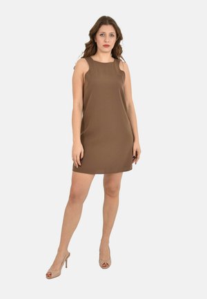 Jonge vrouw met lang bruin haar, gekleed in een mouwloos bruin mini-jurkje en beige hoge hakken, staand tegen een witte achtergrond.